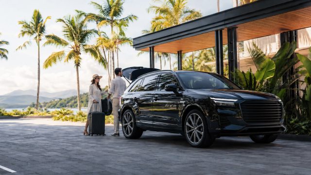 Luxury travellers with luggage beside a premium black SUV at a tropical resort-style arrival area near palm trees and coastal scenery.
