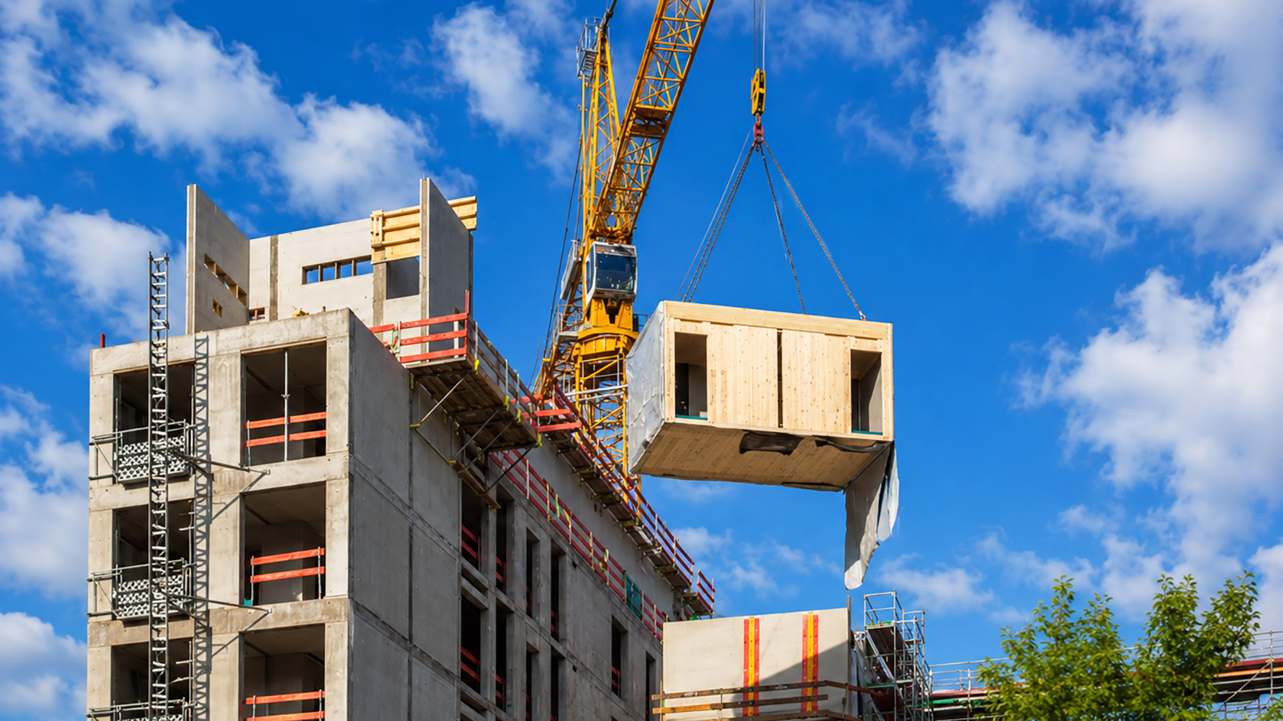 A tower crane lifts a prefabricated timber module into place beside a multi-storey commercial building under construction.