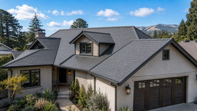 Northern California home with a clean slate roof, straight ridge line, beige stucco exterior, dark trim, wood garage doors, drought-tolerant landscaping, trees, hills, and blue sky.