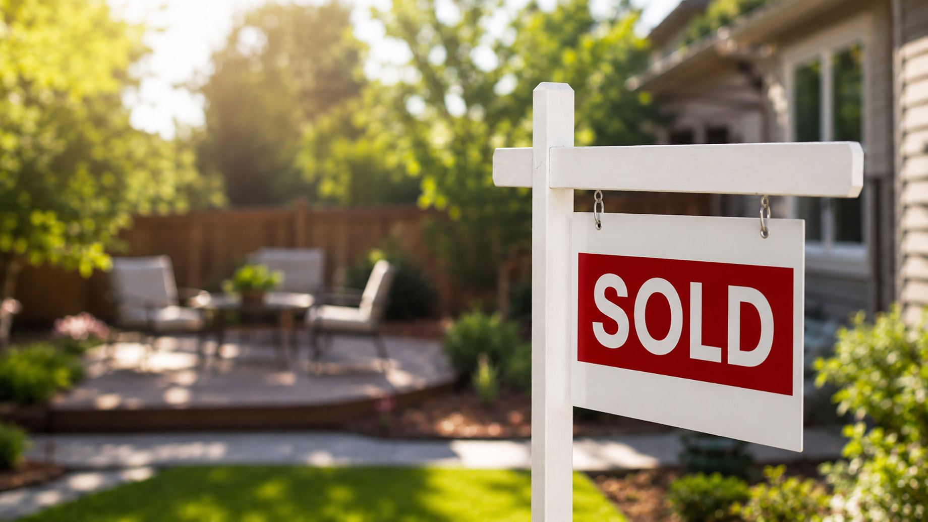 White real estate sign with red sold lettering in front of a bright suburban backyard with patio seating, greenery, fencing, and soft sunlight.