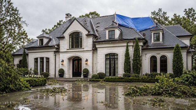 Elegant mansion after severe weather with a blue tarp covering part of the roof, scattered branches on a wet driveway, storm clouds, and landscaped grounds.