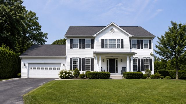 A white two-story suburban house with black shutters, a front lawn, attached garage, and surrounding greenery on a clear day.