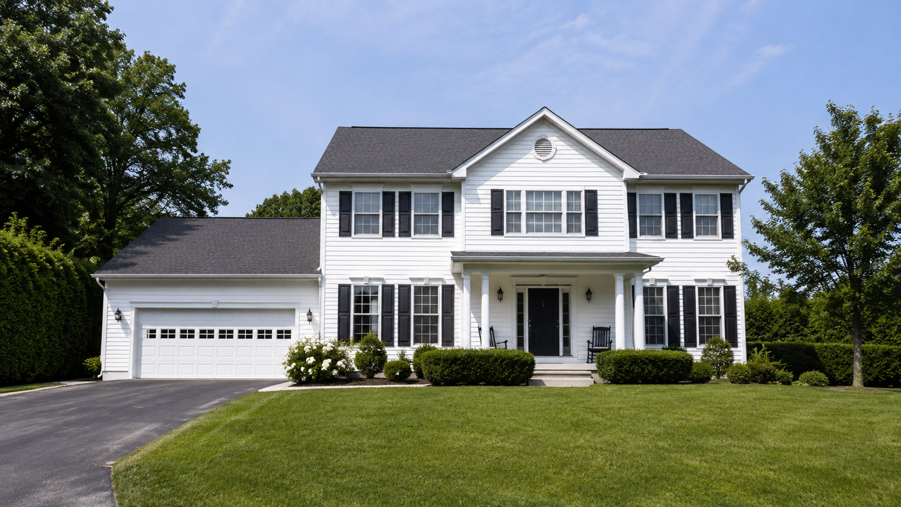 A white two-story suburban house with black shutters, a front lawn, attached garage, and surrounding greenery on a clear day.