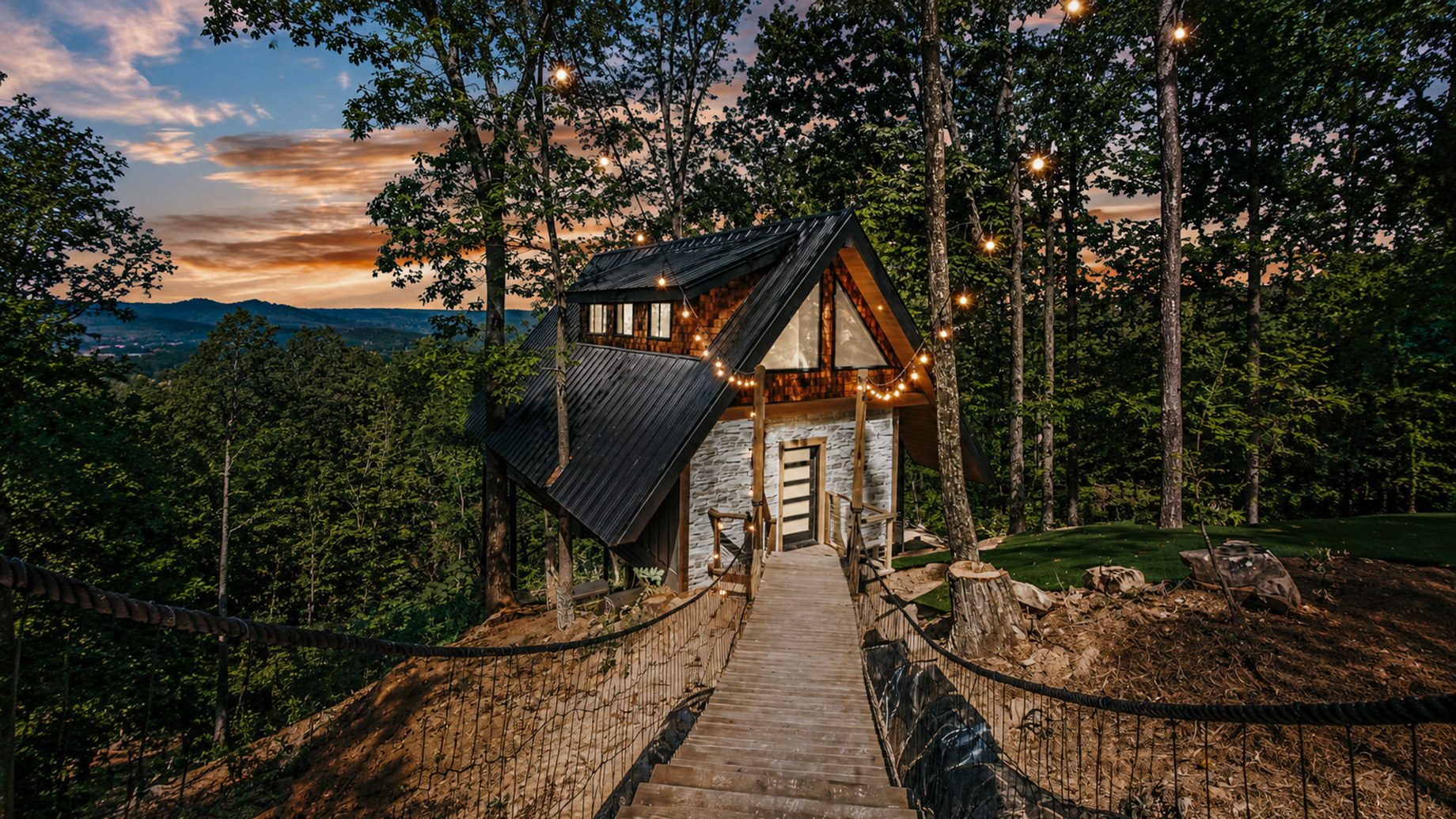A secluded wood-and-stone cabin in a forest setting at dusk, reached by a rope bridge and surrounded by tall trees with warm string lights glowing overhead.