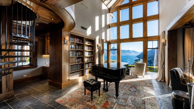 Luxury living room with a grand piano, tall wood-framed windows, built-in bookshelves, vaulted ceiling, patterned rug, and expansive lake and mountain views.