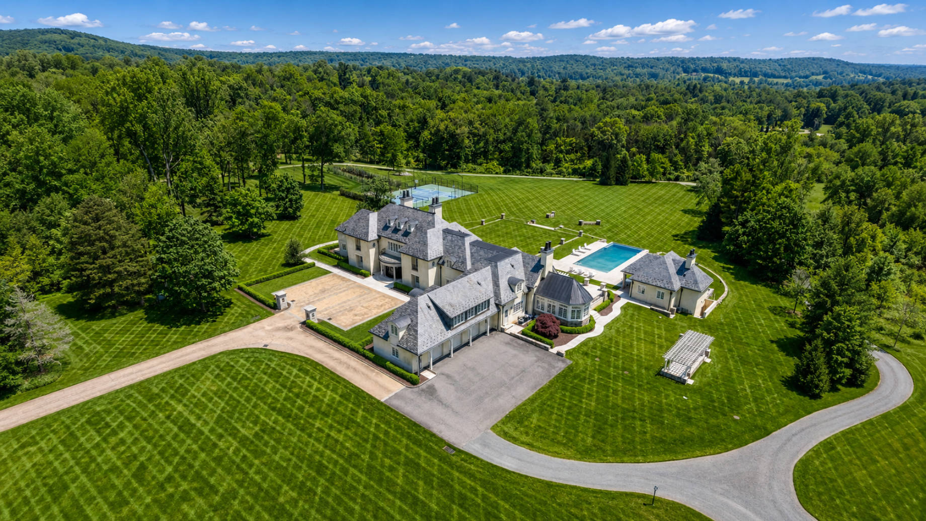 Aerial view of an expansive luxury estate with multiple rooflines, chimneys, a swimming pool, tennis court, and landscaped grounds.