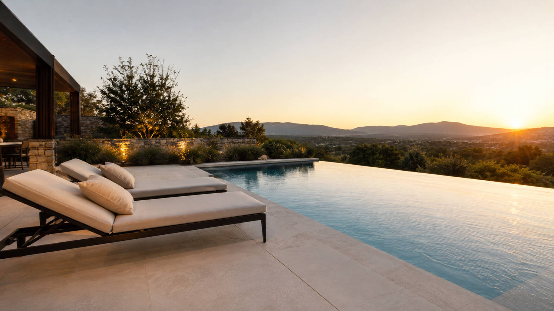 Modern luxury home terrace with an infinity pool, lounge chairs, stonework, and sunset views over a green hillside.