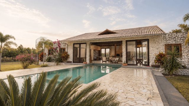 Luxury stone villa with a swimming pool, tropical landscaping, large glass doors, and a soft sunset sky over an outdoor living terrace.