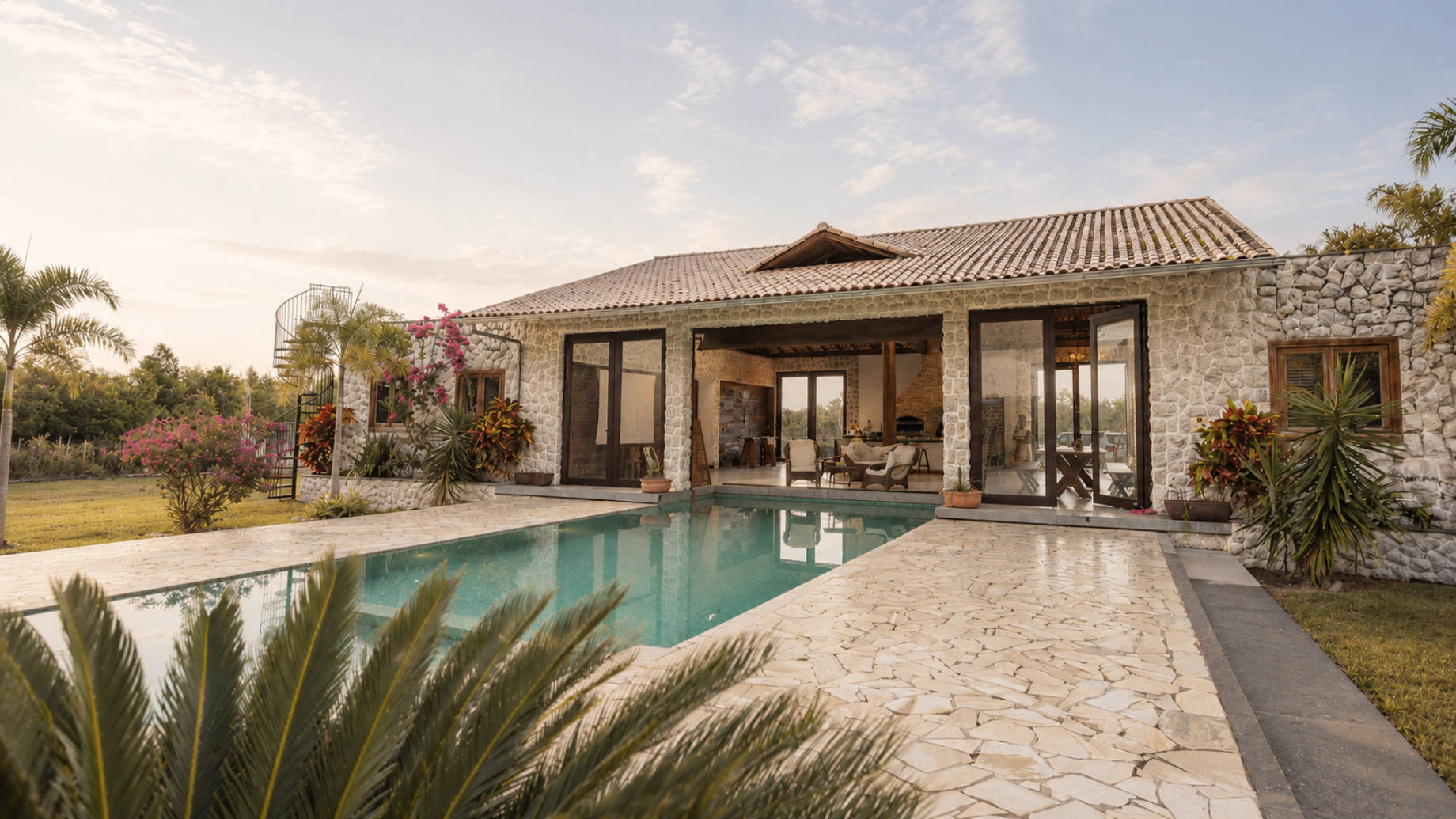 Luxury stone villa with a swimming pool, tropical landscaping, large glass doors, and a soft sunset sky over an outdoor living terrace.