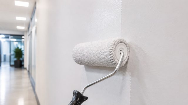Close-up of a paint roller applying a fresh white finish to a commercial hallway wall with a clean, modern corridor in the background.