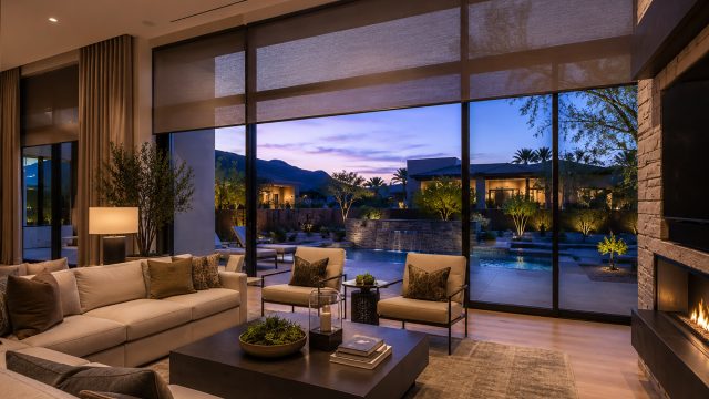 Modern luxury home interior with large glass doors, soft window coverings, warm evening light, and a private pool terrace beyond.