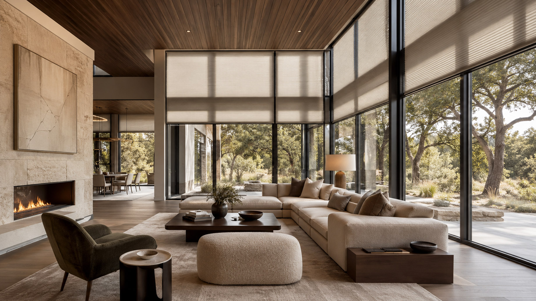 Luxury modern living room with floor-to-ceiling glass, partially lowered cellular shades, warm wood ceilings, stone fireplace, neutral furnishings, and views of mature trees.