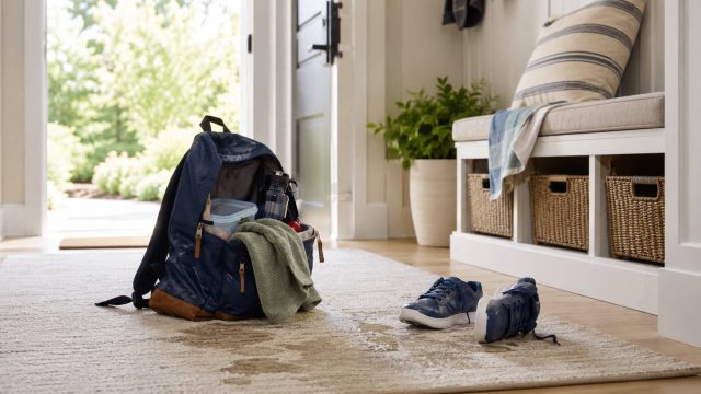 Damp summer camp backpack, wet towel, shoes, and small water marks on a light rug inside a bright home entryway.