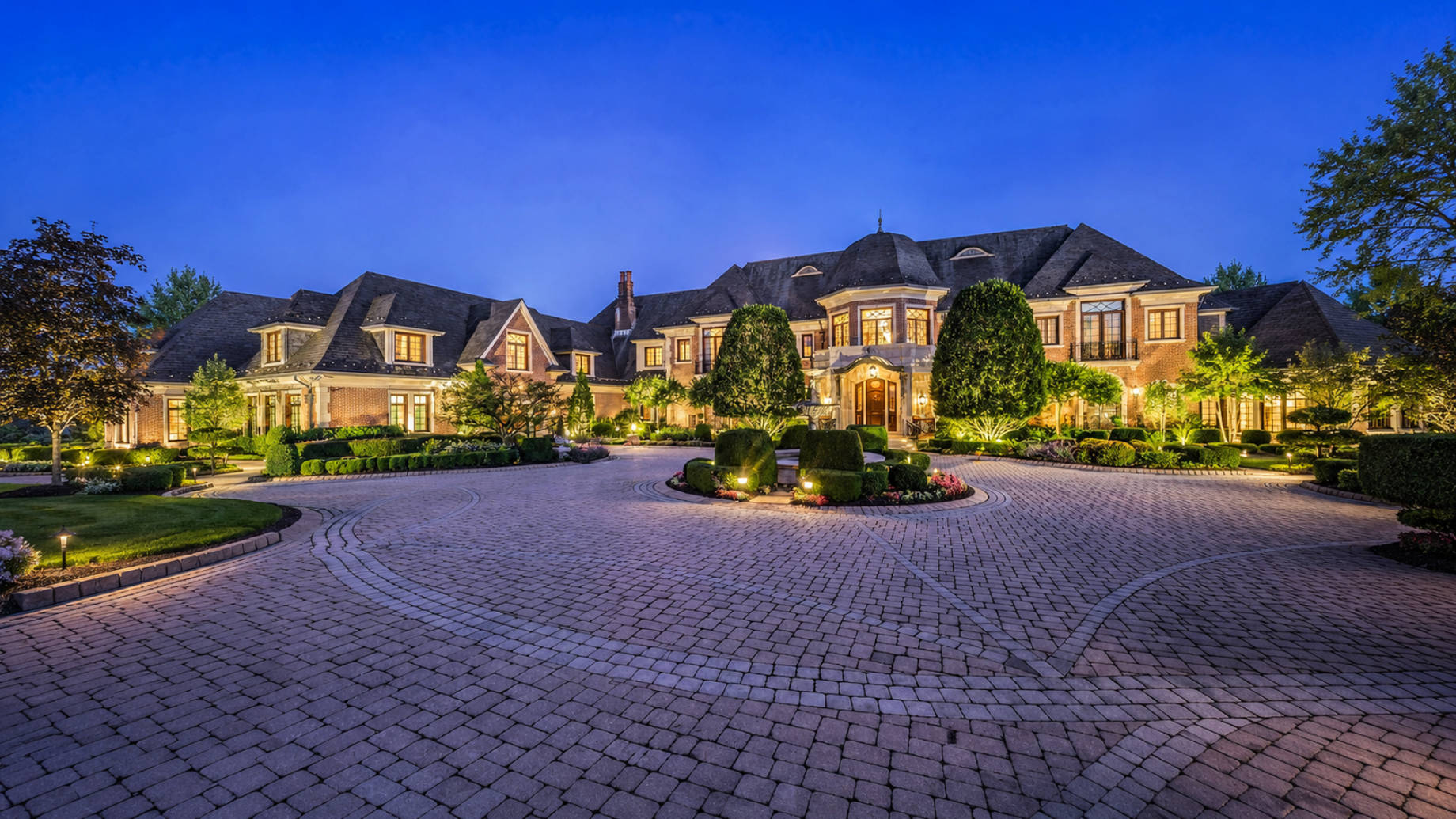 Grand brick mansion at twilight with a dark slate roof, illuminated entrance, manicured gardens, and a circular stone driveway.
