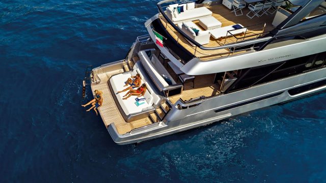 Overhead view of a modern yacht aft deck with sun pads, lounge seating, teak decking, and swimmers beside deep blue water.