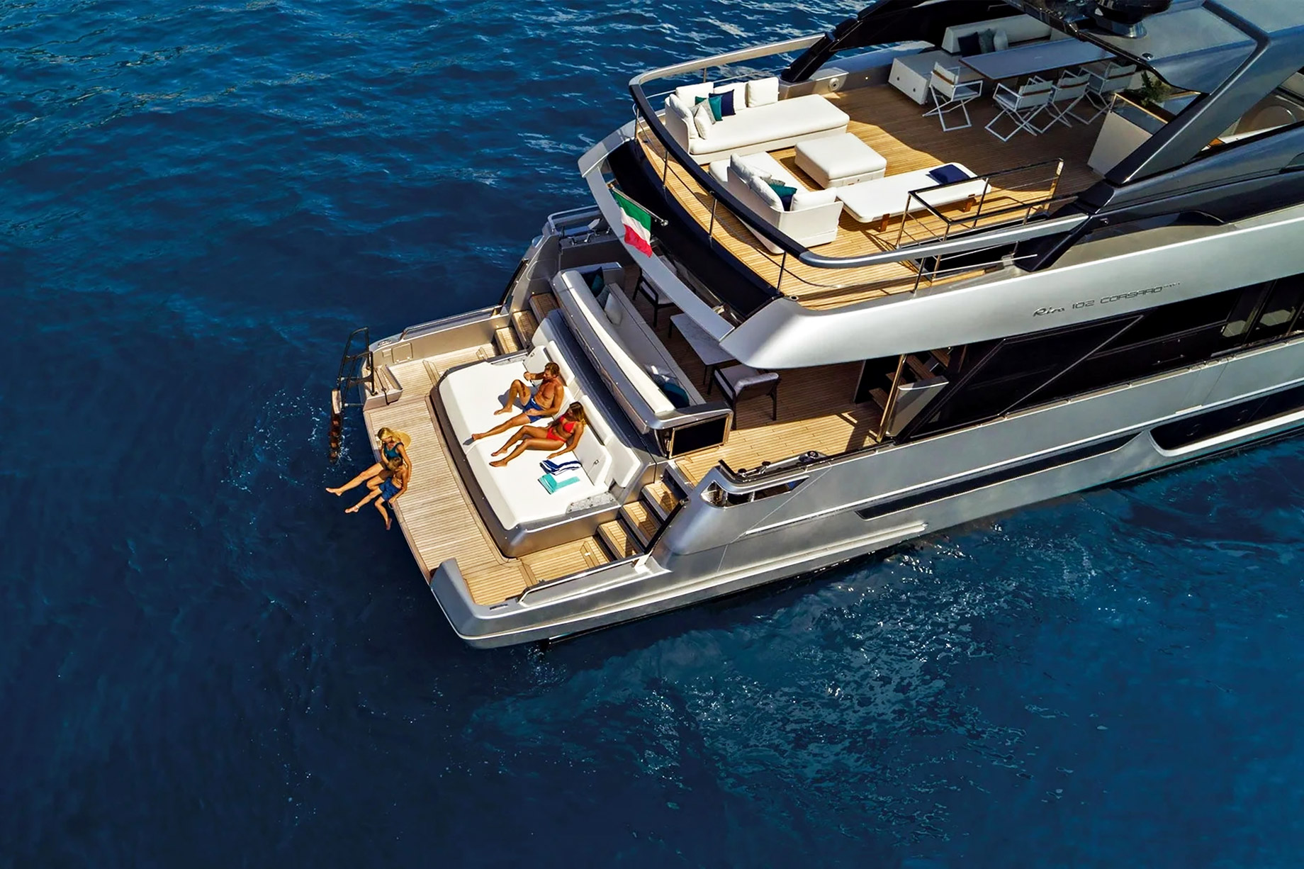 Overhead view of a modern yacht aft deck with sun pads, lounge seating, teak decking, and swimmers beside deep blue water.