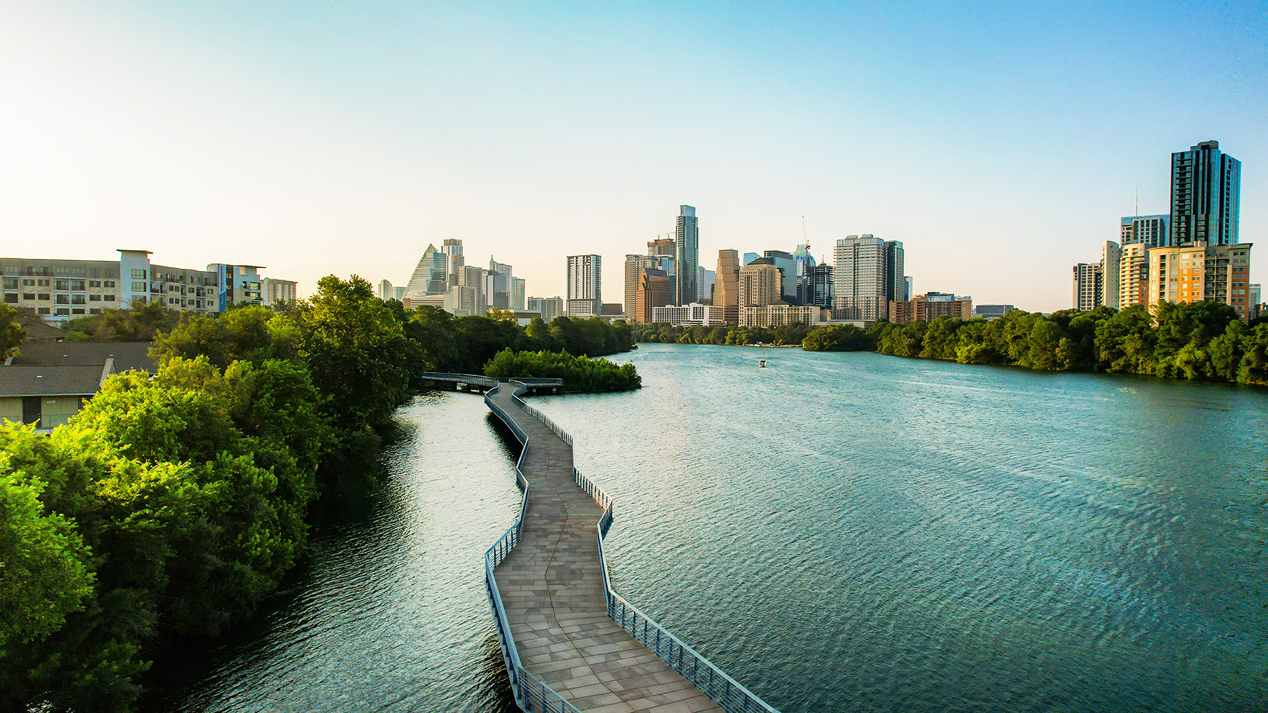 Austin skyline behind Lady Bird Lake with a waterfront boardwalk, green trees, high-rise buildings, and apartment towers.