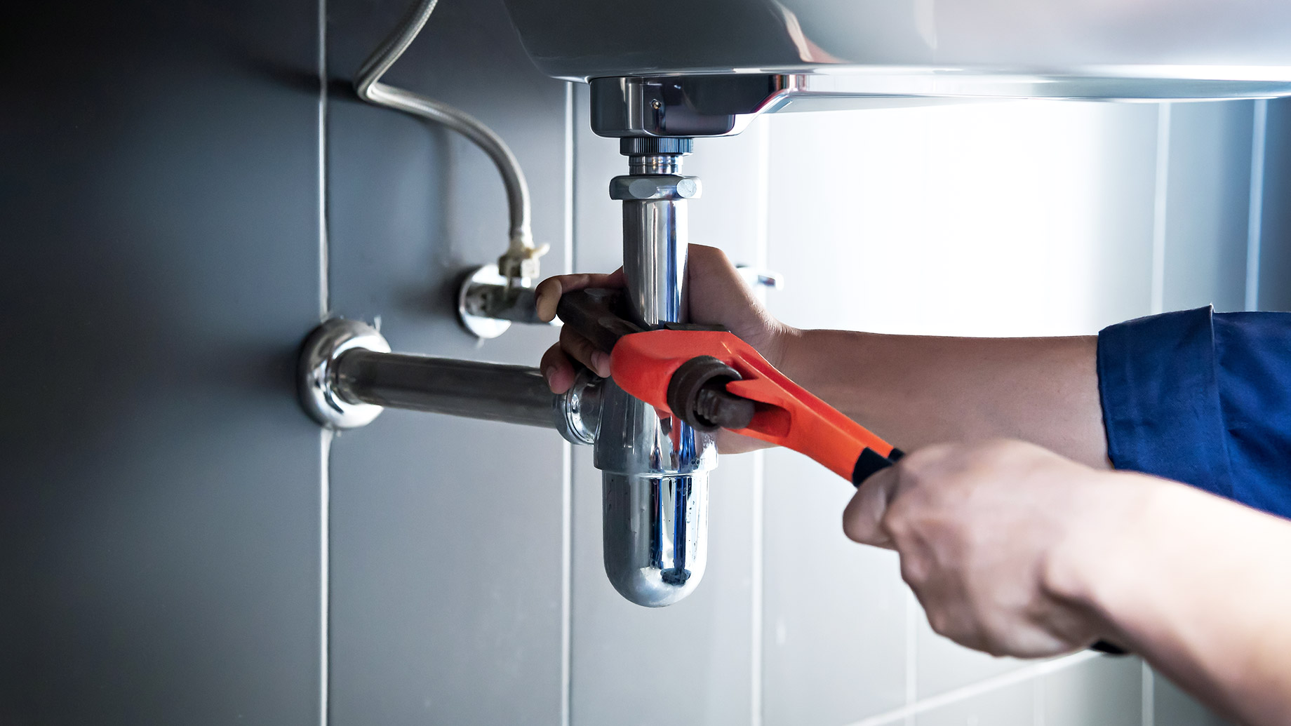 Plumber using a pipe wrench to repair chrome pipework beneath a bathroom sink.
