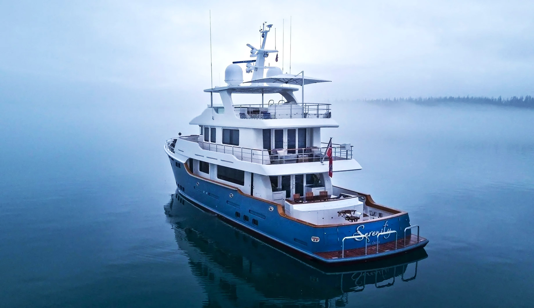 Blue-and-white motor yacht floating on calm, fog-covered water with a faint forested shoreline in the distance.