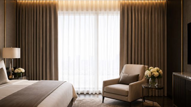 Hotel-inspired bedroom with layered sheer curtains and heavy blackout drapes, warm recessed lighting, neutral bedding, an armchair, and elegant side table.