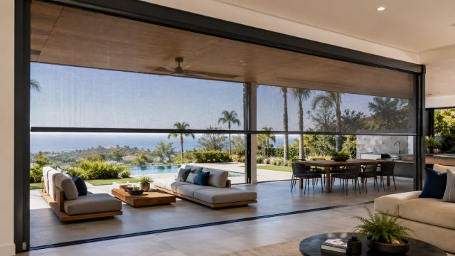 Large-span motorized patio screens enclosing a luxury Australian alfresco area with outdoor seating, dining space, pool, and coastal views.