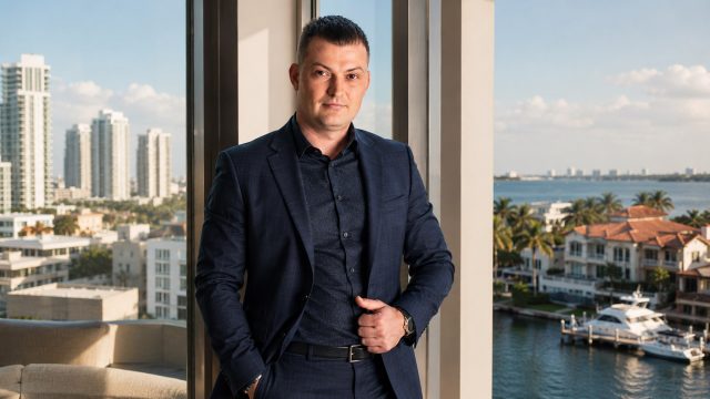 Maksym Herasimov standing in a luxury waterfront residence overlooking high-rise buildings, coastal homes, palm trees, and yachts in Florida.