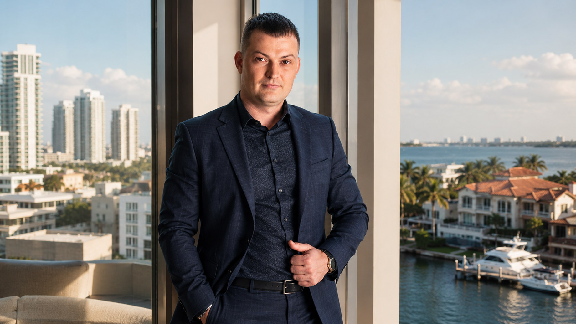 Maksym Herasimov standing in a luxury waterfront residence overlooking high-rise buildings, coastal homes, palm trees, and yachts in Florida.