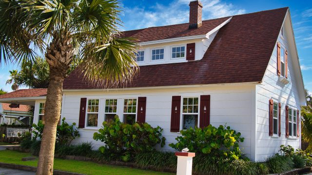 White coastal home in Melbourne, Florida with a red shingle roof, palm trees, shutters, and bright curb appeal under a blue sky.