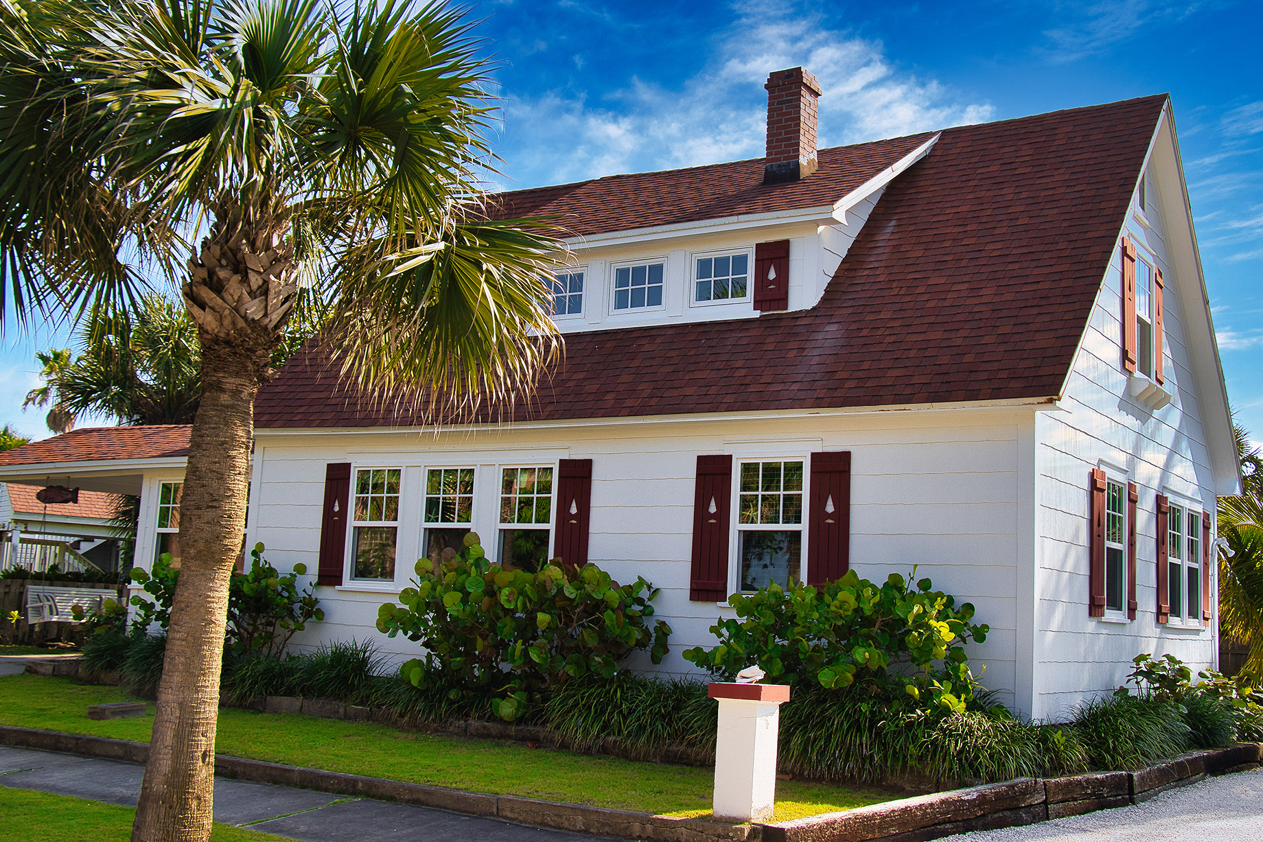 White coastal home in Melbourne, Florida with a red shingle roof, palm trees, shutters, and bright curb appeal under a blue sky.