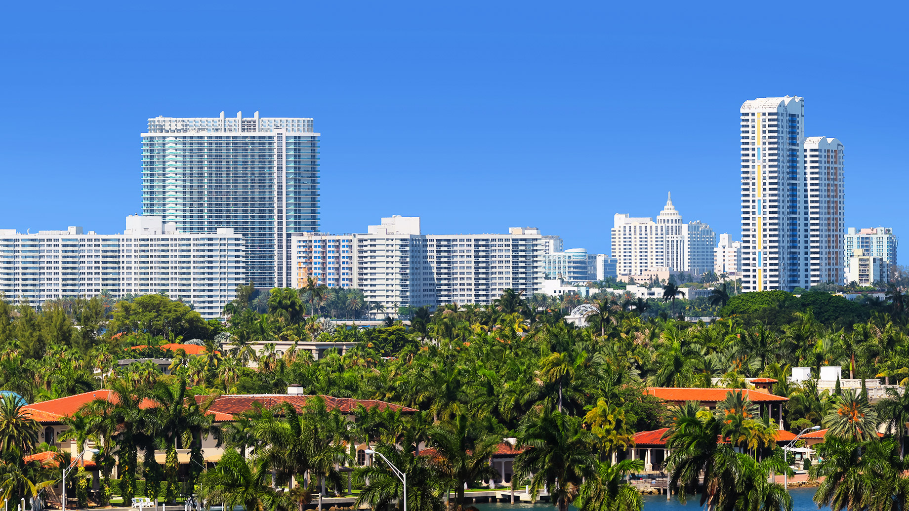 Miami shoreline with luxury homes, palm trees, high-rise apartments, and condominium towers under a clear blue sky.