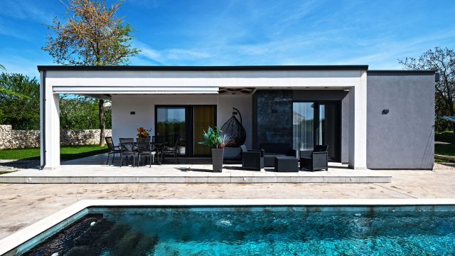 Single-story modern prefab-style home with a covered terrace, outdoor dining area, lounge seating, large glass doors, and a private swimming pool.