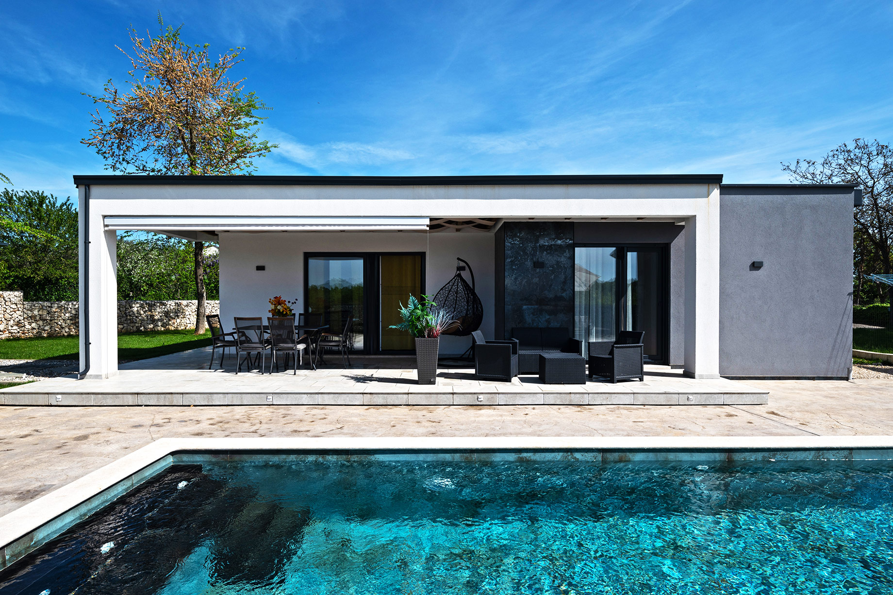 Single-story modern prefab-style home with a covered terrace, outdoor dining area, lounge seating, large glass doors, and a private swimming pool.