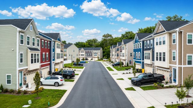Modern townhomes along a clean suburban street with driveways, parked cars, small lawns, sidewalks, and a bright blue sky.