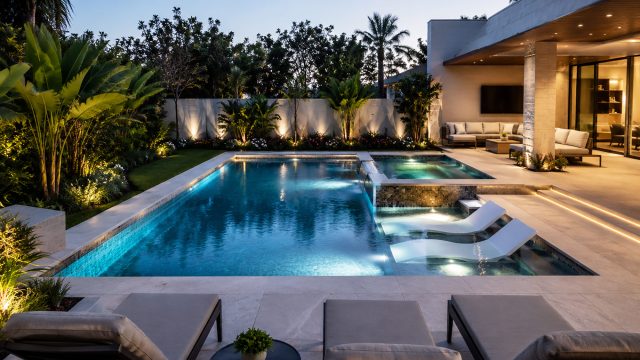 Renovated luxury swimming pool with contemporary stone paving, integrated lighting, a raised spa, landscaped greenery, and a modern outdoor lounge.