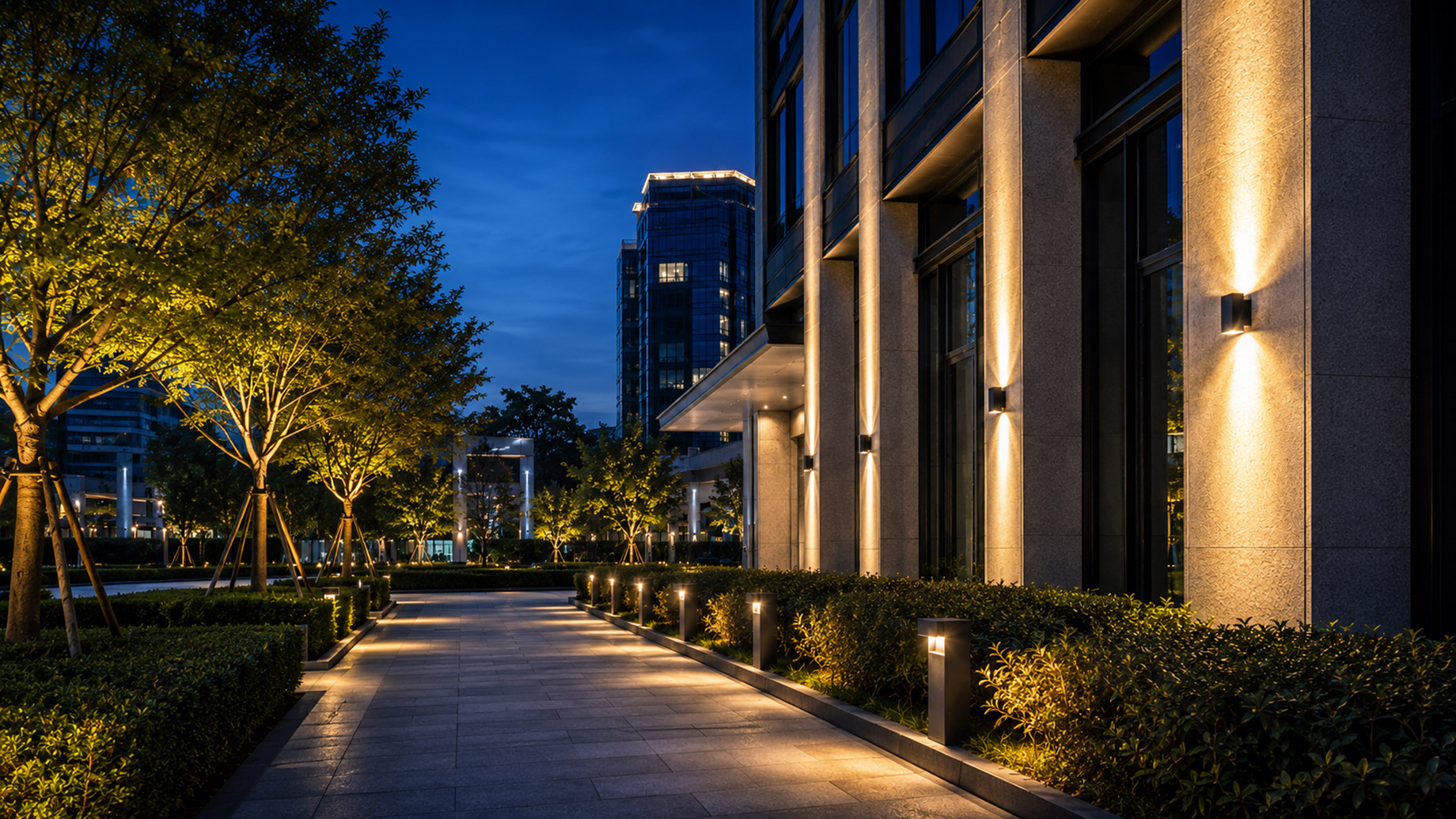 Commercial building and landscaped pedestrian walkway illuminated by architectural wall lights, pathway bollards, and integrated outdoor LED fixtures at dusk.
