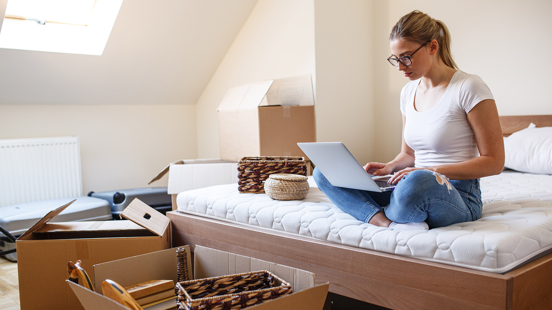Woman sitting on a mattress with a laptop while packing boxes and baskets in a bright bedroom during a one-bedroom move.