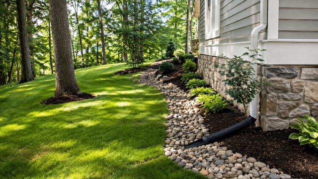 Graded lawn beside a home with a downspout extension directing water into a stone-lined drainage channel away from the foundation.