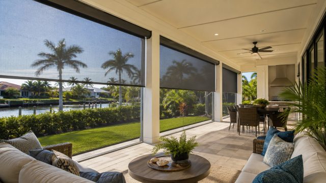 Covered Florida patio with lowered retractable screens, outdoor seating, dining area, palm trees, and waterfront views.