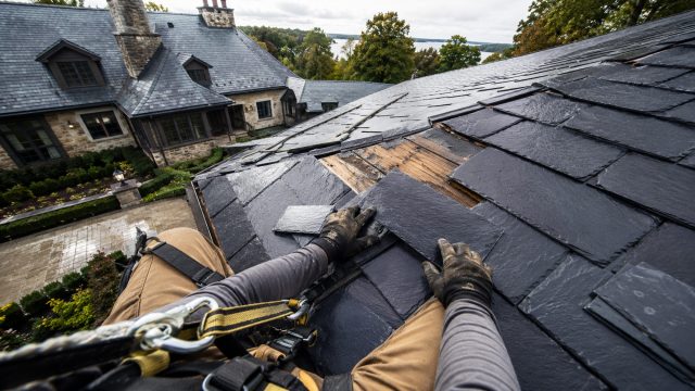 Roofer wearing a secured safety harness reinstalls slate tiles over a storm-damaged section of a luxury stone home’s roof.
