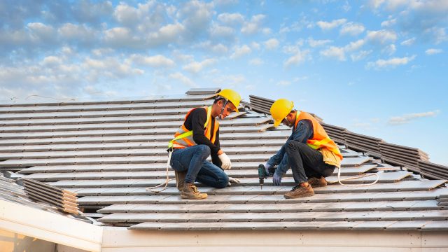 Two roofers wearing hard hats and safety harnesses installing roofing materials on a residential home under a bright sky.