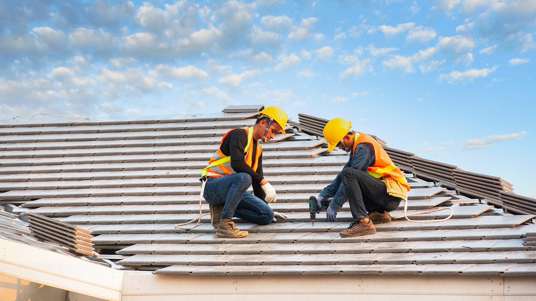 Two roofers wearing hard hats and safety harnesses installing roofing materials on a residential home under a bright sky.