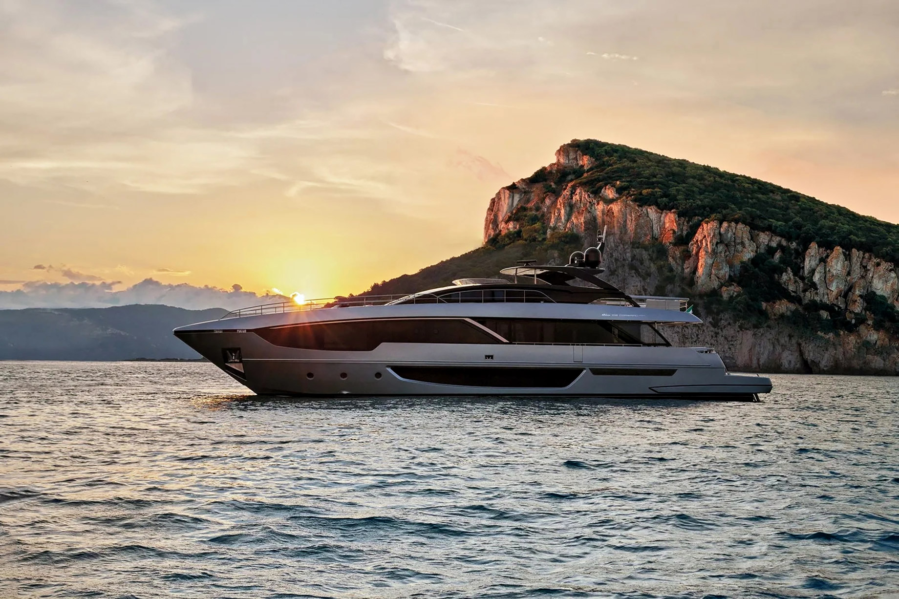 Low-profile luxury motor yacht on open water at sunset with a rocky coastline in the background.