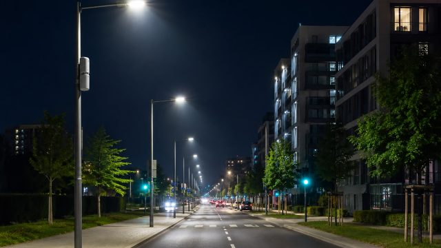 Modern city road at night with illuminated smart streetlights, mounted control devices, apartment buildings, trees, and light traffic.