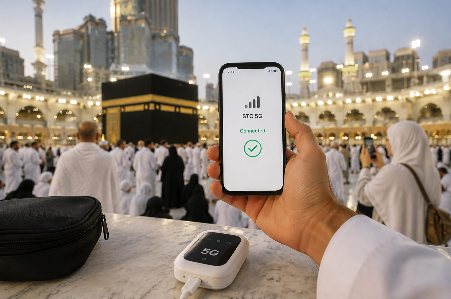 Hand holding a smartphone showing a 5G connection near the Kaaba at Masjid al-Haram, with pilgrims and a portable mobile hotspot.