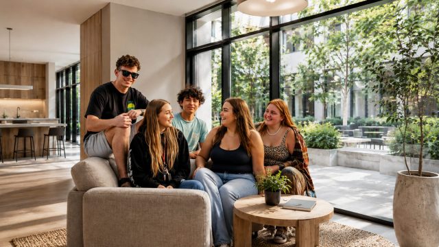 Five university students relaxing together in a bright modern lounge overlooking a landscaped courtyard.