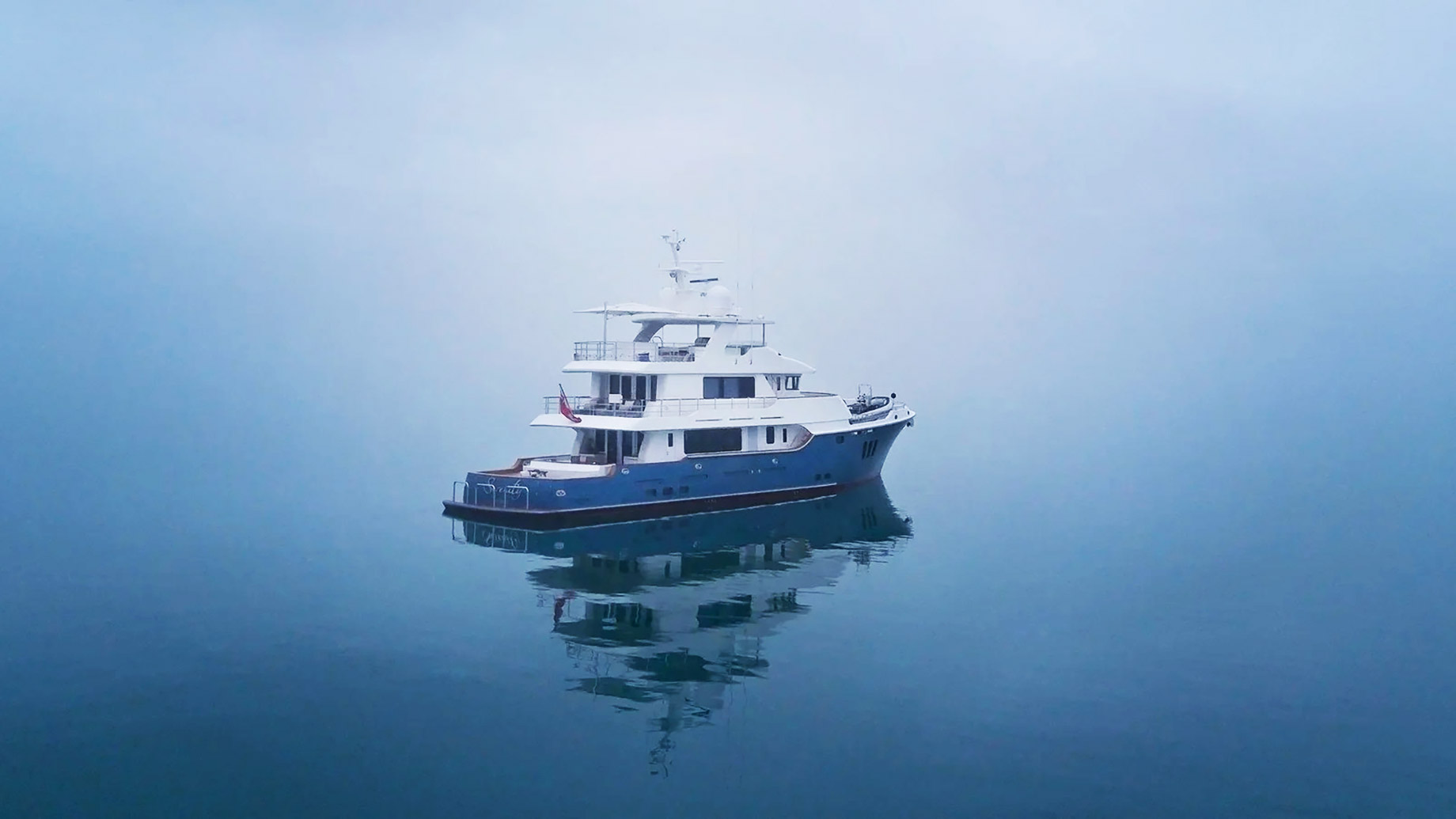 White-and-blue motor yacht anchored in dense fog on calm water with a soft reflection beneath the hull.