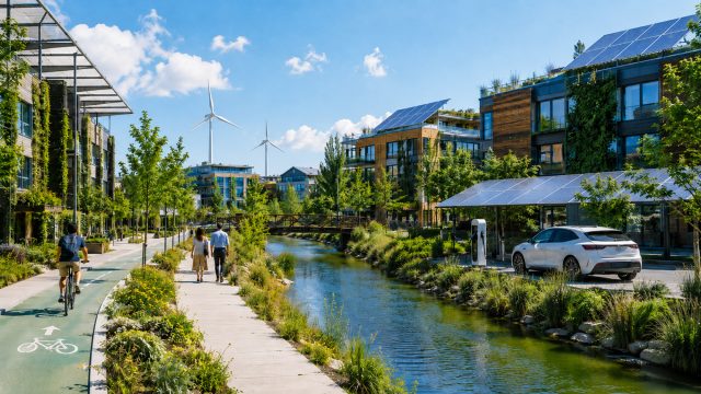 Modern eco-friendly riverside district with solar panels, wind turbines, green buildings, an EV charging station, bike lane, pedestrians, and native landscaping.