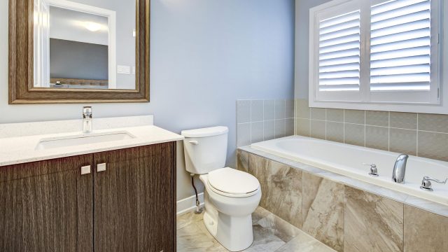 Bright bathroom with a wood vanity, stone-look flooring, white soaking tub, toilet, tiled surround, and plantation shutters.