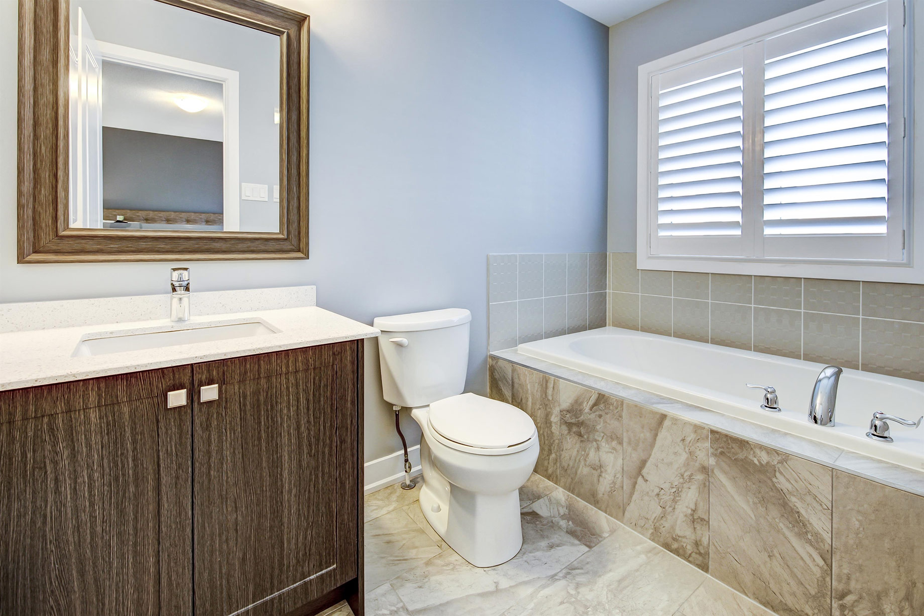 Bright bathroom with a wood vanity, stone-look flooring, white soaking tub, toilet, tiled surround, and plantation shutters.