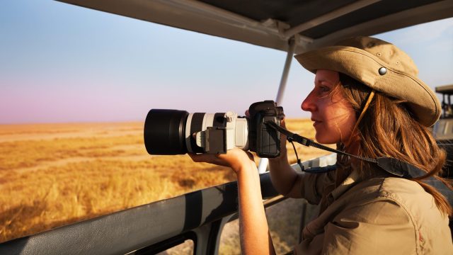 Woman holding a professional telephoto camera aboard an open safari vehicle overlooking the sunlit African savannah.